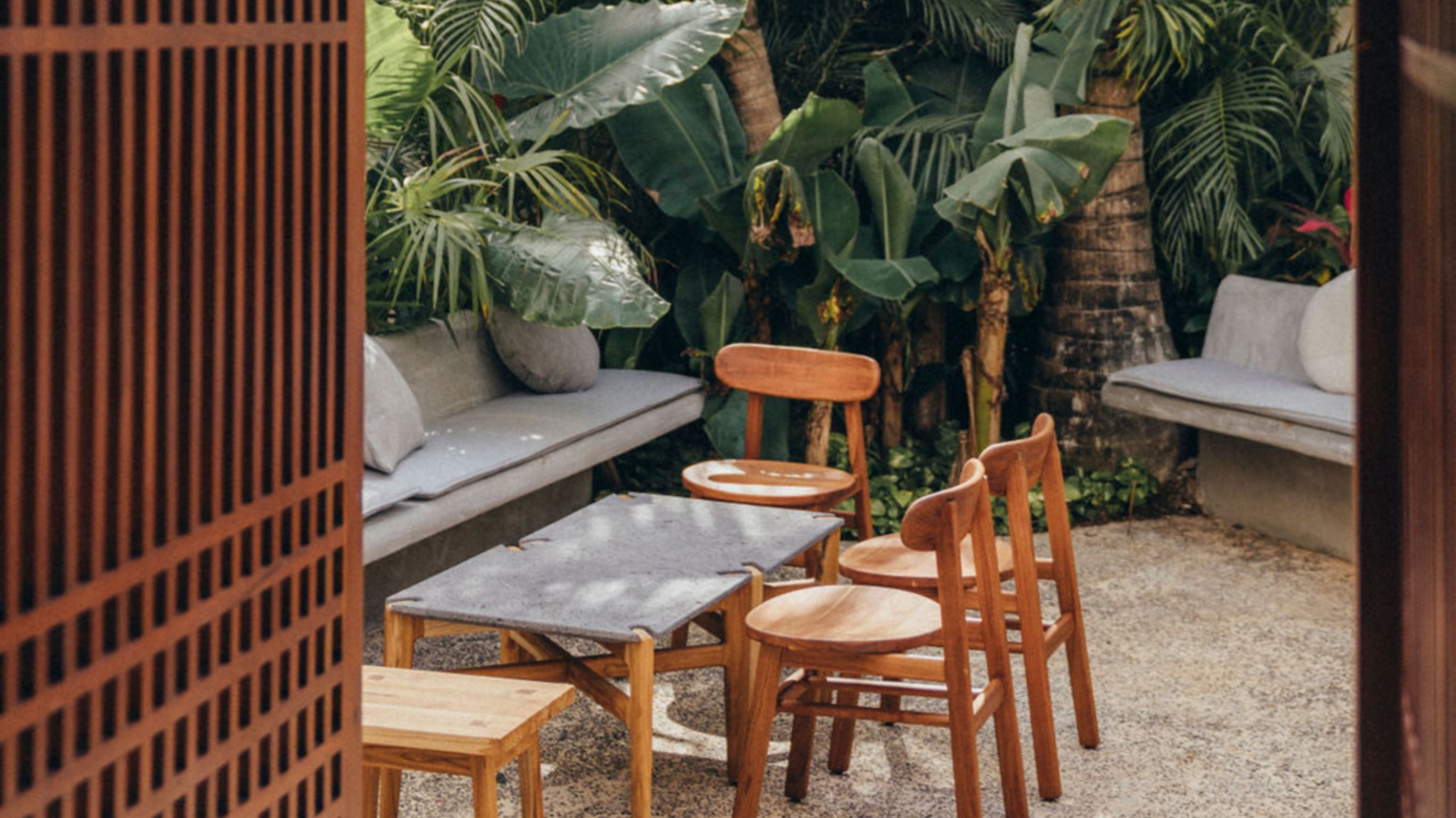 Vista de sillas de madera y una pequeña mesa afuera sobre concreto con la vegetación